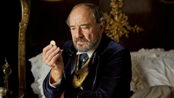 Der alte Uhrmachermeister, gespielt von Viktor Preiss, sitzt auf einem Bett und betrachtet gedankenversunken eine Goldmünze, die er vor sich in der Hand hält.