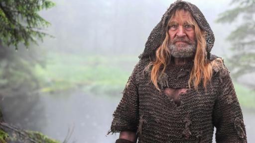 Vor dem Hintergrund eines Waldsees, der in eine Wiese ausläuft steht der Eisenhans (Michael Mendl). Er trägt Bart, eine weite Kapuze über dem Kopf, hat lange, blonde Haare und trägt ein schmutzig-braun wirkendes Gewand.