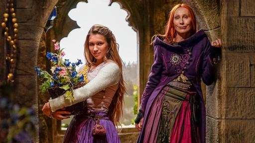 In einem alten, steinernen Gemäuer stehen Rapunzel, gespielt von Anne-Lena Schwing, und Zauberin Eleonor, gespielt von Andrea Sawatzi, vor einem steinernen Fensterrahmen und schauen in die Kamera.
