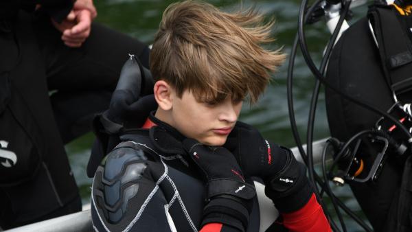 Der Junge Freddy sitzt an der Rehling eines Bootes und hat einen Neoprenanzug und Handschuhe an.