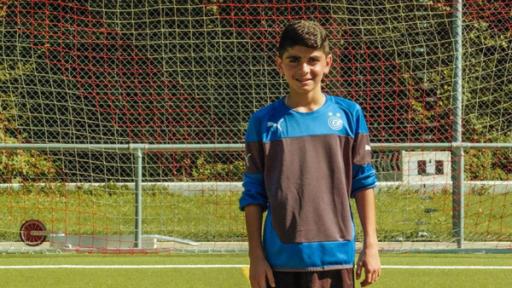 Ayham steht auf dem Fußballplatz vor dem Tor. Er trägt ein blau/schwarzes Trikot und schaut freundlich in die Kamera. Die Sonne scheint.