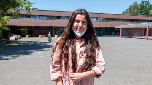 Azra steht mit heruntergezogener Maske vor dem Schulgebäude. Azra hat langes dunkles Haar, trägt eine Jeans und eine rosafarbene Jacke. Der Schulhof hinter ihr ist leer. Sie lacht in die Kamera.