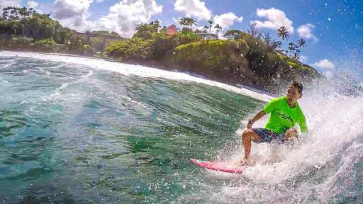 Diesel - Die perfekte Welle | Rechte: ZDF Diesel trägt ein neonfarbenes Surfshirt, dunkle Surfshorts und surft eine Welle. Das Wasser spritzt hinter ihm auf. Im Hintergrund ist das Ufer zu erkennen.