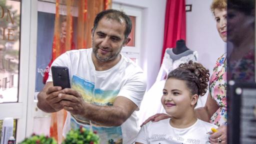 Fetije - Mein Leben bestimme ich | Rechte: ZDF Fetije sitzt geschminkt und frisch frisiert im Salon und lächelt, dabei schaut sie in das Handy ihres Vaters, der neben ihr steht und ein Foto von ihr macht. Die Stylistin steht hinter ihr und hat die Hände auf Fetijes Schultern gelegt. Sie ist nur im Anschnitt zu sehen. Im Hintergrund sieht man ein Brautkleid auf einer Büste.