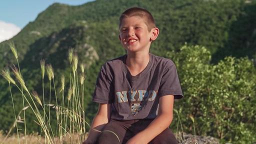 Ilija - Mein erster Wettlauf | Rechte: ZDF Ilija sitzt auf einem Felsen und lächelt fröhlich in die Kamera. Die Sonne scheint. Im Hintergrund kann man einen bewaldeten Hang erkennen. Alles wirkt sehr ruhig und friedlich.