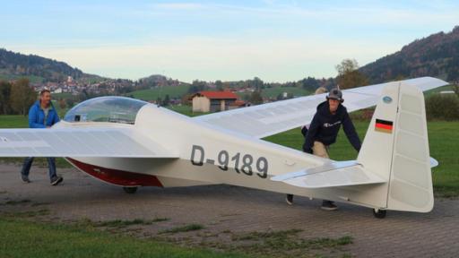 Marius - Allein durch die Lüfte | Rechte: ZDF Marius bringt zusammen mit seinem Fluglehrer seinen Flieger zur Startbahn. Sein Trainer schiebt den vordereren Teil des Flugzeugs, während Marius das Heck anschiebt. Im Hintergrund ist eine Berglandschaft zu sehen.