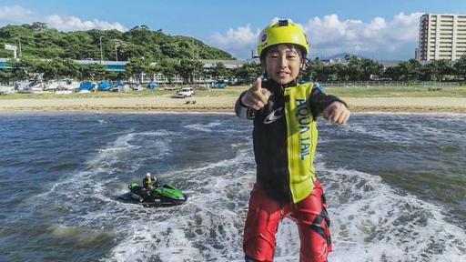 Yushi befindet sich in der Luft über dem Wasser. Er trägt seine Wassersportbekleidung und einen gelben Schutzhelm. Er zeigt mit seiner rechten Hand den Daumen nach oben und lächelt in die Kamera. Im Hintergrund sind der Strand und Hochhäuser zu sehen.