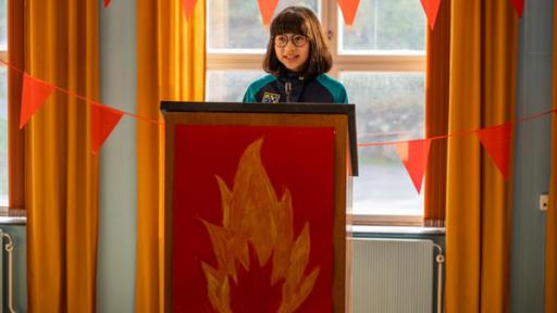 Ein fast fairer Wahlkampf | Rechte: © SEEFOOD TV AS 2023/Eline Ødegaard Alpha steht lächelnd und nach vorne blickend an einem Rednerpult, auf dem eine Flamme abgebildet ist. Direkt vor ihr befindet sich ein Mikrofon. Hinter ihr hängen orangefarbene Wimpelketten am Fenster.