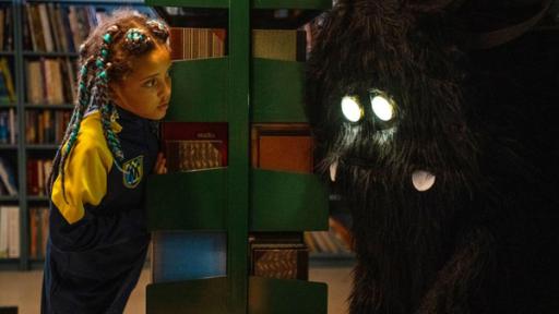 Immer schön bescheiden bleiben | Rechte: © SEEFOOD TV AS 2023/Eline Ødegaard In der Bibliothek der Schule schaut Jenny an ein Bücherregal gelehnt und mit ungläubigem Blick auf ein zotteliges Wesen mit leuchtenden Augen, das vor ihr steht.
