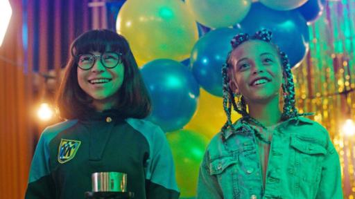 Neue Schüler und alte Freunde | Rechte: © SEEFOOD TV AS 2023/Eirik Evjen Die beiden Mädchen Alpha und Jenny stehen glücklich lächelnd und nach vorne blickend nebeneinander in der Schulaula. Im Hintergrund hängen blaue und gelbe Luftballons als Partydekoration.