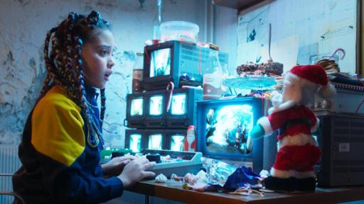 Weihnachten allein in der Schule | Rechte: © SEEFOOD TV AS 2023/Mads Sæther Juul Jenny steht mit erschrockenem Blick an einem Tisch und schaut auf einen von vielen kleinen Monitoren einer Überwachungskamera, die sich vor ihr befinden. Direkt vor ihr steht ein Bedienpult mit verschiedenen Knöpfen. Auf dem Tisch befinden sich zudem verschiedene, teils ausgepackte, weihnachtliche Süßigkeiten sowie eine Nikolaus-Stofffigur.
