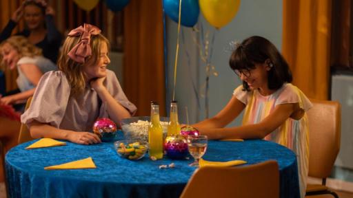 Wenn Turteltauben tanzen gehen | Rechte: © SEEFOOD TV AS 2023/Eline Ødegaard Aurora und Alpha sitzen gemeinsam an einem runden Tisch mit blauer Samt-Tischdecke. Auf dem Tisch stehen zwei Limonadenflaschen, bunte Becher mit Strohhalmen, sowie gelbe Servietten, eine große Glasschüssel mit Popcorn und eine kleine Schüssel mit diversen Süßigkeiten. Aurora schaut Richtung Alpha und lächelt. Diese grinst. Beide Mädchen tragen ihre bunten Ballkleider. Im Hintergrund hängen zwei Luftballons.