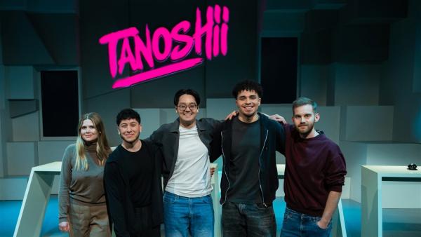 Im Tanoshii-Studio stehen von links nach rechts:. Creatorin Closetanimegirl, Umut vom Kanal Kyodai, Moderator Raafey, Luis vom Kanal Kyodai und Creator JayDred. Sie stehen eng nebeneinander in der Mitte des Studios und lächeln. Im Hintergrund ist die Studiokulisse mit Pulten und oben mittig das Tanoshii-Logo zu sehen.