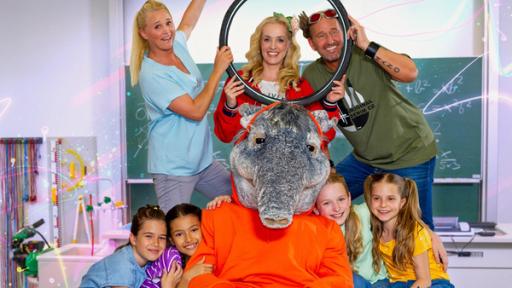 Rechte: KiKA/ZDF/Ron Bergmann Moderatorin Singa, Kindermusikerin Saskia Süß und Tom Lehel halten lachend vor einer Schultafel einen Fahrradreifen hoch. Die TanzAlarm-Kids sitzen gemeinsam mit TanzTapir davor.