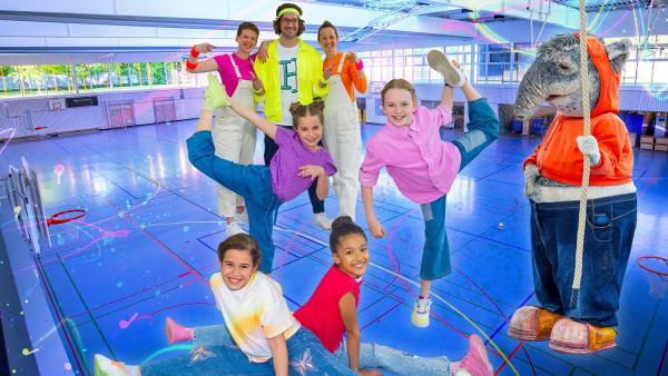 Die TanzAlarm-Kids stehen in sportlichen Posen vor Kindermusiker herrH und seinen Tänzerinnen mitten in der Sporthalle. TanzTapir steht rechts an der Seite mit einem Kletterseil in der Hand.