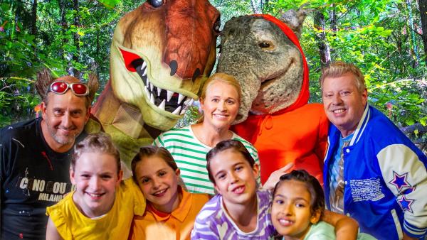 Tom Lehel, ein T-Rex, TanzTapir, Singa, Kindermusiker Volker Rosin und die TanzAlarm-Kids stehen mitten im Wald unfd lächeln in die Kamera.
