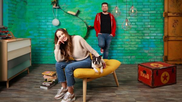 Im Team Timster-Studio: Soraya sitzt auf einem gelben gepolsterten Sessel. Neben ihr liegt Hund Jimmy. Tim steht dahinter vor einer türkise Ziegelwand. Beide lächeln.