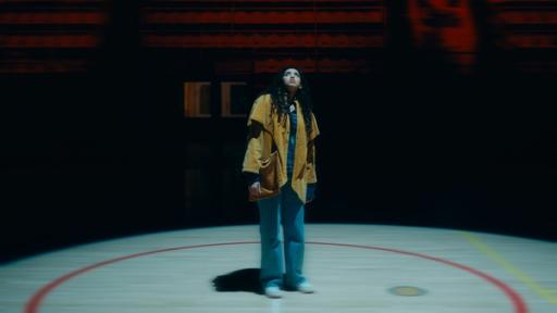 Der Stich | Rechte: KIKA Hawa steht allein in einer Turnhalle. Ein Spot ist von oben auf sie gerichtet. Sie schaut ängstlich nach oben. Die Stimmung ist surreal.