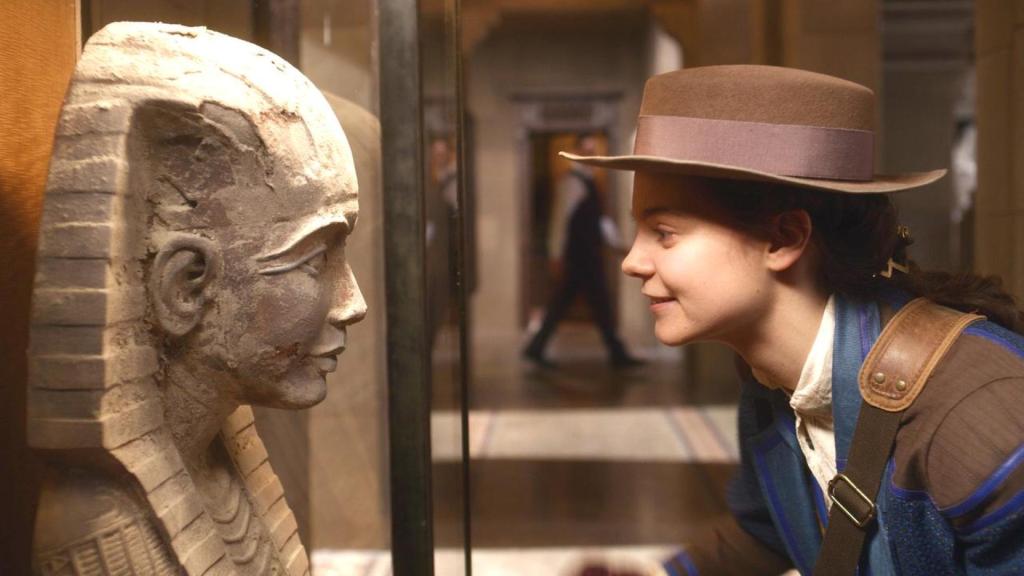 Im Museum: Theodosia (Eloise Little) schaut lächelnd und leicht zu ihr hin gebeugt, einer steinernen Pharaonen-Statue in die Augen. Sie trägt einen braunen Hut.