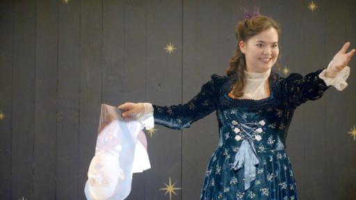 Josie steht auf der Bühne und zaubert mit echter Magie. Sie hat die linke Hand zum Publikum ausgestreckt und in der rechten eine zusammengefaltete Papierfigur von Theo.