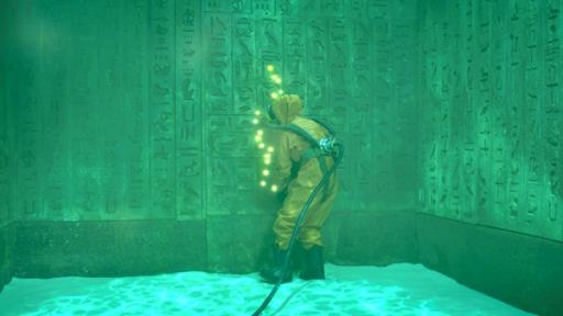 Unter Wasser, in einer gefluteten Kammer, umgeben von Wänden voller Hieroglyphen mit dem Rücken zum Bild: Theo, die eine Tauchausrüstung trägt, steht vor einer dieser Wände. Gelb leuchtende Punkte erscheinen vor ihr und bilden ein senkrecht, kurviges Muster.