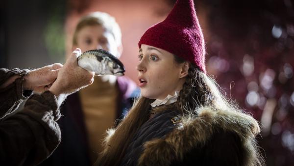 Tinka seitlich mit Wichtelmütze. Mit weit geöffneten Augen starrt sie auf einen Fisch, der ihr in zwei Händen vor die Nase gehalten wird.