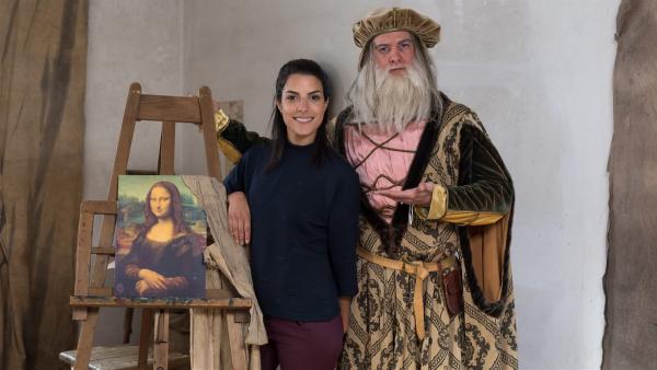 Clarissa steht neben Leonardo da Vinci (Philipp Danne). Er hält mit einer Hand die Staffelei und mit der anderen zeigt er auf Clarissa.