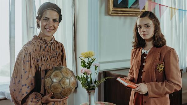 Clarissa trifft auf Anne Frank (Katharina Kron). Clarissa lächelt und hat einen alten Fussball in der Hand. Anne Frank schaut neutral und hält ein Buch in ihren Händne.
