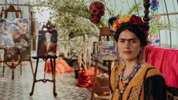 Die mexikanische Malerin Frida Kahlo (Mina Sagdic) bei ihrer Vernissage. Im Hintergrund sind Gemälde von ihr zu sehen.