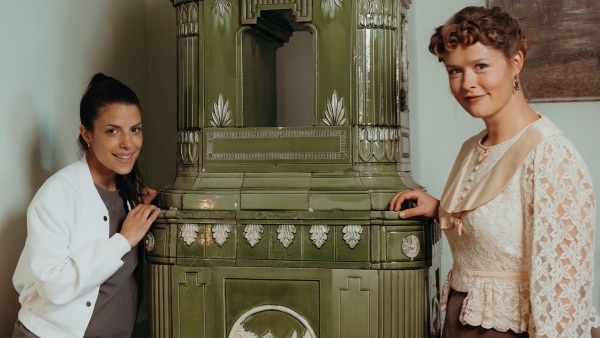 Clarissa und Bertha stehen links und rechts neben einem großen Kachelofen in einem Zimmer und lächeln. Links ist ein kleiner Tisch mit Büchern und einer Lampe. An den Wänden sind zwei Gemälde.