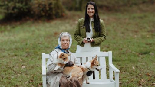 Rechte: KiKA/Sabine Finger Königin Elisabeth II. sitzt mit ihren zwei Corgis auf einer Parkbank. Clarissa steht mit ihrer Zeitmaschine dahinter und lächelt.