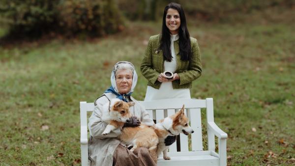 Königin Elisabeth II. sitzt mit ihren zwei Corgis auf einer Parkbank. Clarissa steht mit ihrer Zeitmaschine dahinter und lächelt.