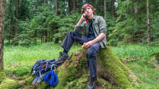 Das Taschenmesser von Rob Rebel | Rechte: ZDF/Mideu Films/Steffen Junghans Mitten im Wald sitzt auf einem Baustumpf der siebzehnjährige Rob. Er trägt ein rotes Stirnband, typische Outdoor-Kleidung, zu seinen Füßen liegt ein Rucksack samt Seil und Kletterhaken. Mit selbstgewisser Mine telefoniert er über sein Handy, hält in seiner anderen Hand ein angebissenes Brötchen und scheint den Moment völlig entspannt zu genießen.