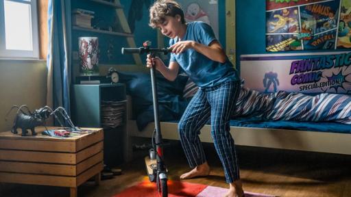 Der coole Scooter | Rechte: ZDF/Mideu Films/Steffen Junghans Jans Zimmer in der ersten Morgensonne: Plakate an den Wänden, im Hintergrund das Bett, links ein Holzschränkchen, auf dem Boden ein bunter Teppich. Darauf steht – im Schlafanzug – Jan (mitte) und umfasst mit beiden Händen, voller Begeisterung, das Lenkrad eines edlen, voll funktionsfähigen und sogar mit einer Lampe ausgestatteten Scooters.
