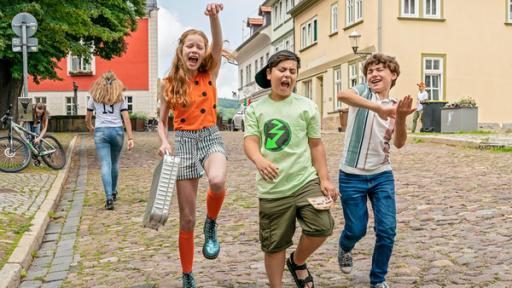 Der Hunderttausend-Euro-Schein | Rechte: ZDF/Mideu Films/Steffen Junghans Auf einer mit Kopfstein gepflasterten, verkehrsberuhigten städtischen Straße kommen uns Patty (v.l.n.r.), Cem und Jan entgegen. Patty trägt einen silbernen Aluminiumkoffer, Cem einen Wettschein. Alle drei scheinen völlig außer Rand und Band, machen fiebrig-übersteigerte Körperbewegungen und scheinen dabei lauthals zu singen.