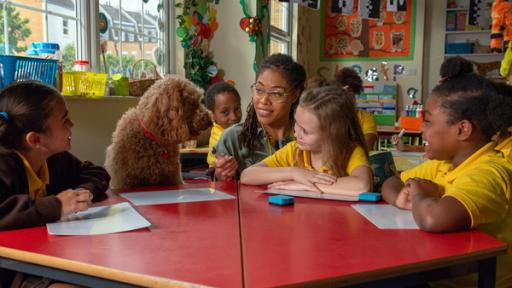 Schreibprobleme | Rechte: ZDF Wuffel sitzt mit Evi und anderen Schülerinnen in der Schule am Tisch. Die Lehrerin kniet neben Wuffel und hält ihm einen Stift hin.