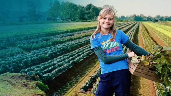 Ein blondes Mädchen steht auf einem Feld und hält eine Holzkiste voll frischem Gemüse im Arm.