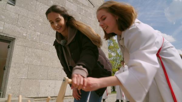 Feiertagsreporterin Soraya und Marlene stehen an einer Kirche und halten gebückt einen Gegenstand aus Holz in den Händen.